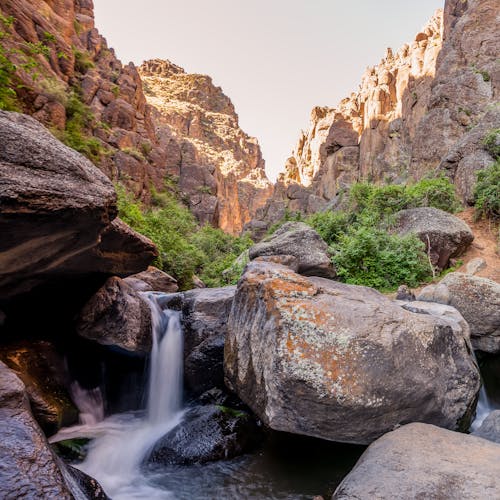 Picturesque scenery of small waterfall flowing through boulders amidst cliffs in breathtaking ravine on sunny day