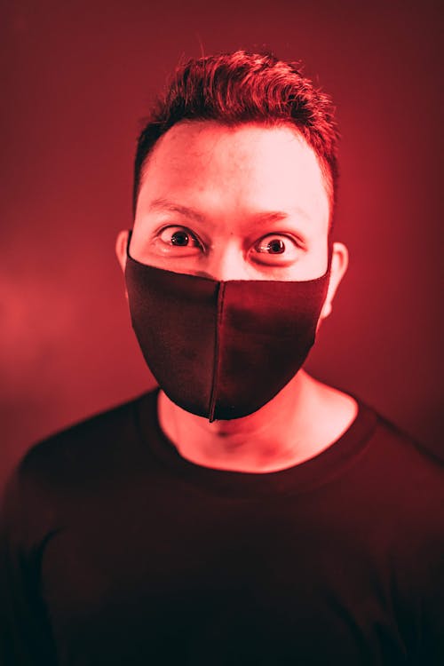 Man with dark hair and bulging eyes wearing black medical mask and sweater in room with red light