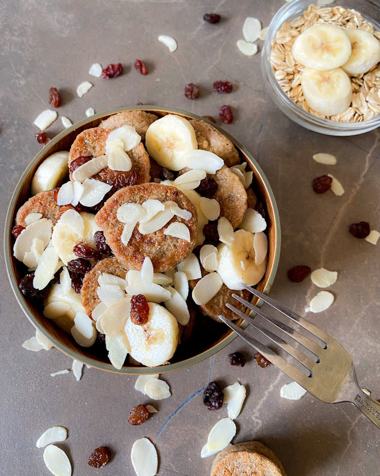 A Bowl Of Mini Pancakes With Almonds, Banana And Raisins On Top