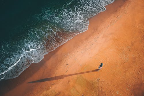 Drone view of person standing on wet sandy beach near foaming waves of ocean