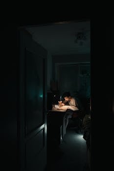A young man studies intensely at his desk at night, illuminated by a desk lamp.