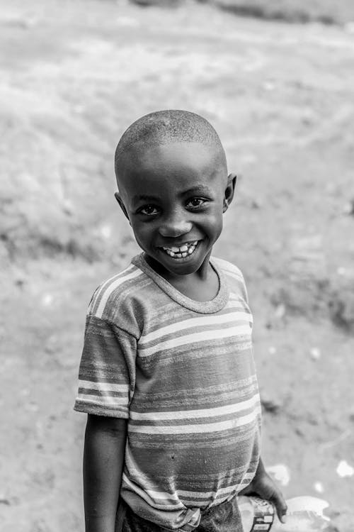 Black and white cute little ethnic boy with toothy smile standing on street of poor village