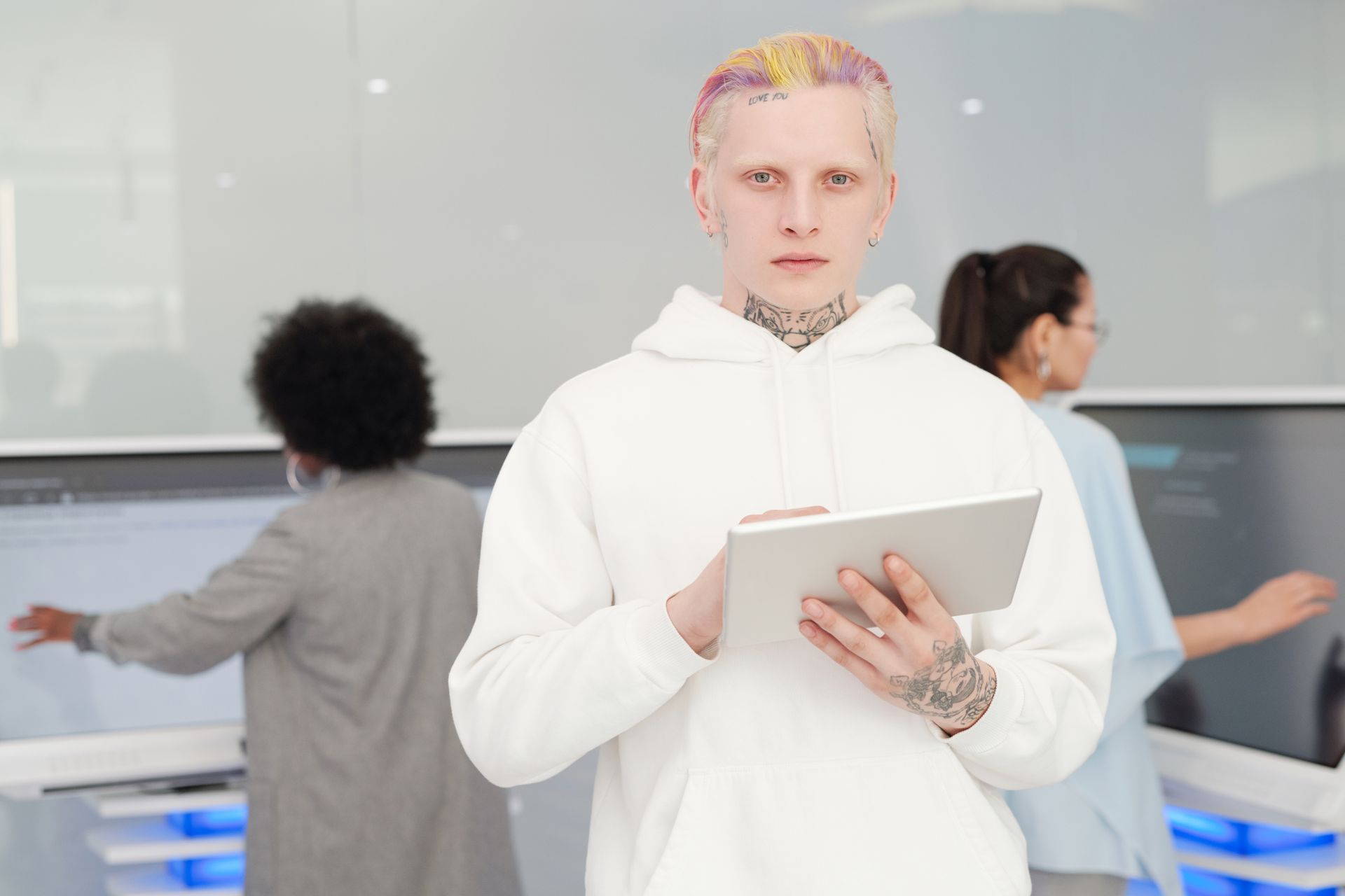 Tattooed man in white hoodie using a tablet in a tech environment with diverse colleagues.