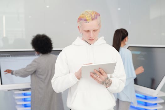 A young man with colorful hair and tattoos using a tablet in a modern tech environment while others work in the background.