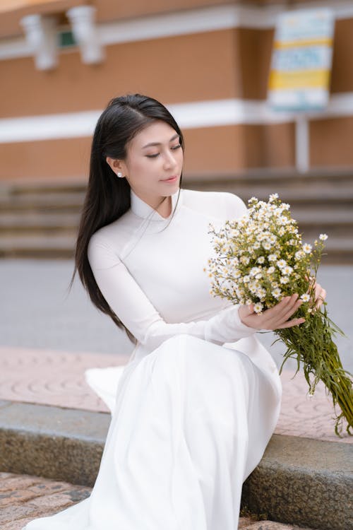 Charming young Asian female wearing white long dress sitting on pavement with bunch of gentle flowers