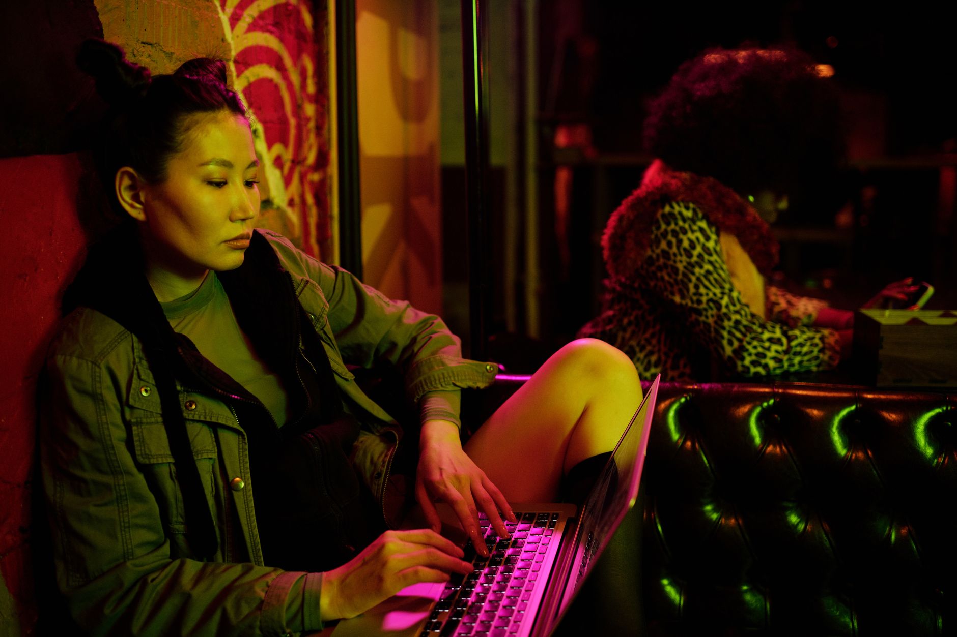 Asian woman working on a laptop in a dimly lit, colorful interior setting, capturing focus and modern lifestyle.