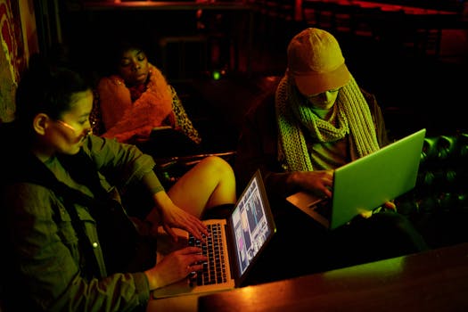 Group of young adults using laptops in cozy, dim setting, conveying cyber atmosphere.