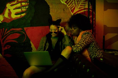 Free Two fashionable women using a laptop in a dimly lit urban cafe with vibrant graffiti walls. Stock Photo