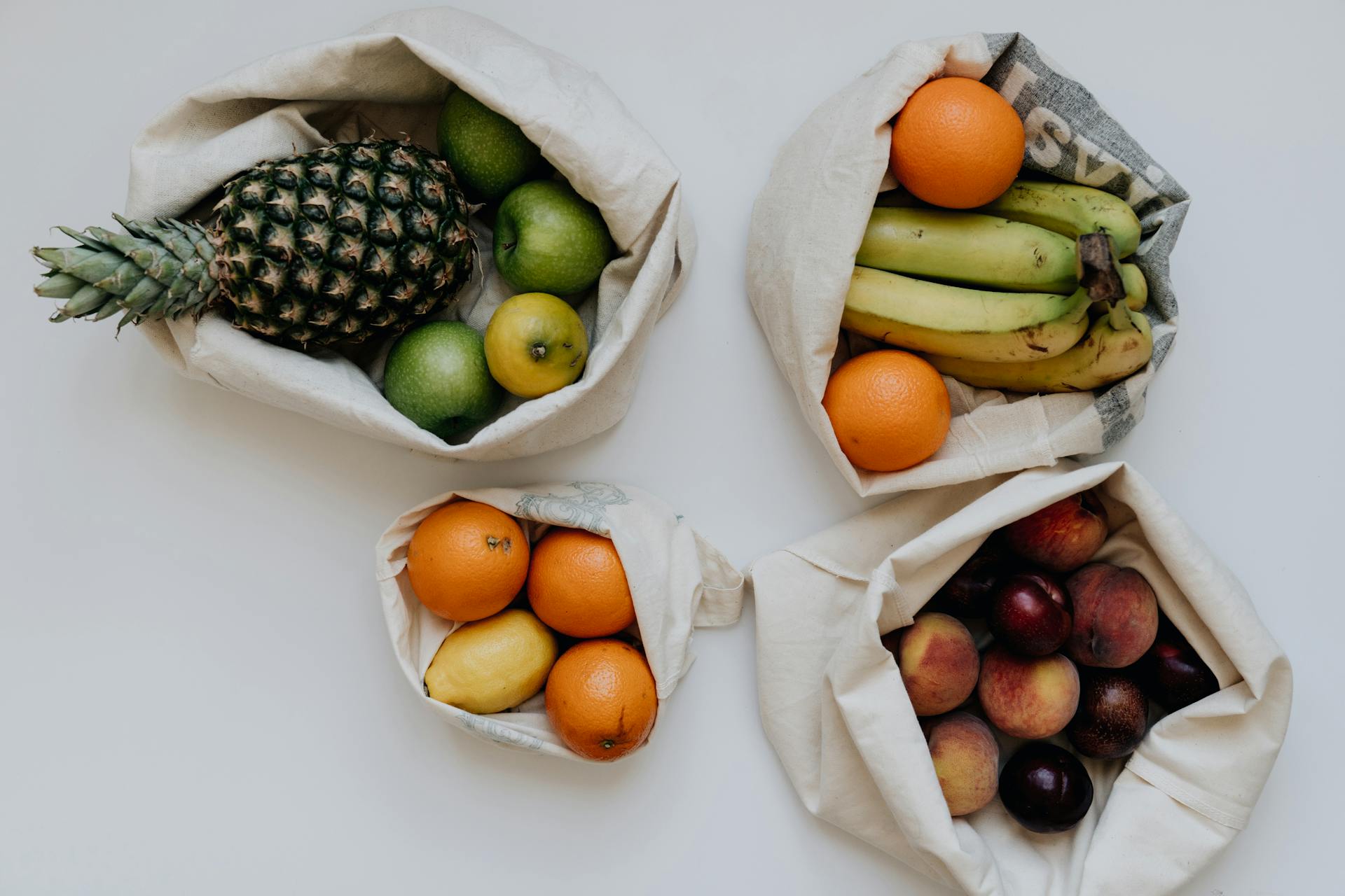 A variety of fresh organic fruits in reusable cotton sacks on a white background for a healthy lifestyle.