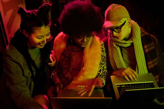 Three diverse adults using laptops, immersed in a collaborative indoor setting with colorful lighting.