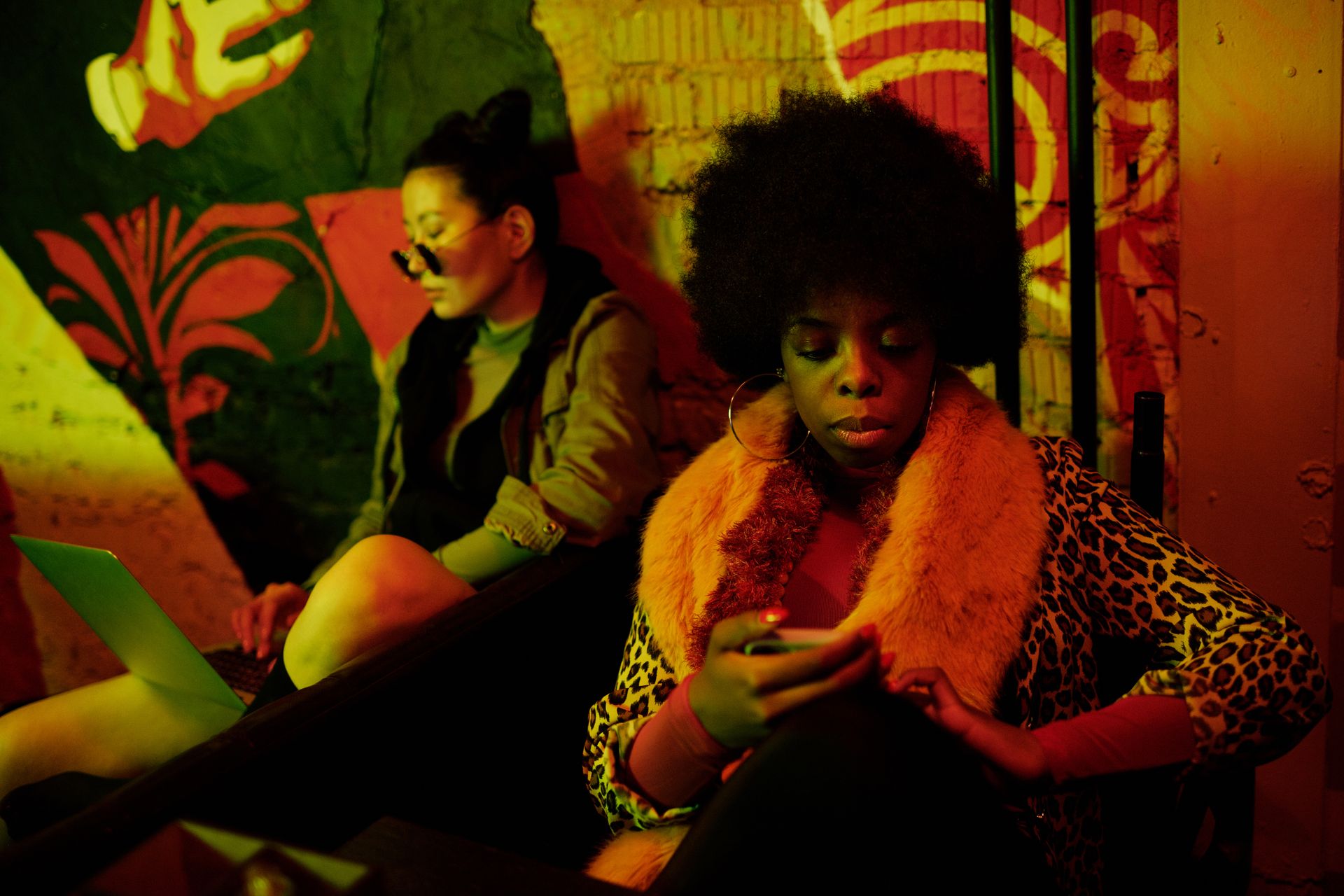 Two women in a vibrant cafe using devices in a creative setting, showcasing diverse fashion and technology use.