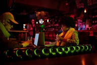 在光线昏暗的酒吧里，两名成年人正用笔记本电脑工作，其中一人手里拿着饮料