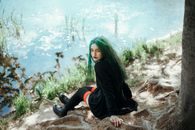 From above full body of pensive adolescent female in black coat and long stockings looking at camera over shoulder while chilling on roots of old tree on coast of lake in sunlight