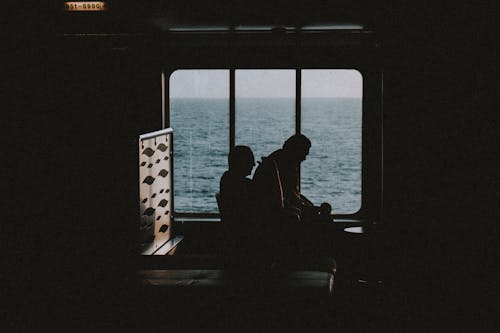 Silhouette of unrecognizable tourists sitting next to window and relaxing in dark room on passenger ship
