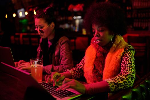 Two women work on laptops in a dimly lit bar, surrounded by vibrant lights and a relaxed atmosphere.
