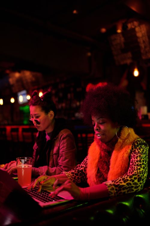 Free Two women typing on laptops in a warmly lit bar, creating a cozy work atmosphere. Stock Photo