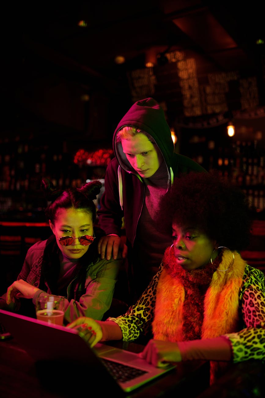 Three young adults collaborate on a project using a laptop in a dimly lit bar.