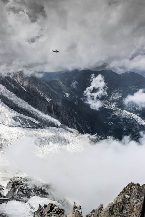 Free Amazing view of helicopter flying above rough mountains peaks covered with snow and fog against cloudy sky Stock Photo