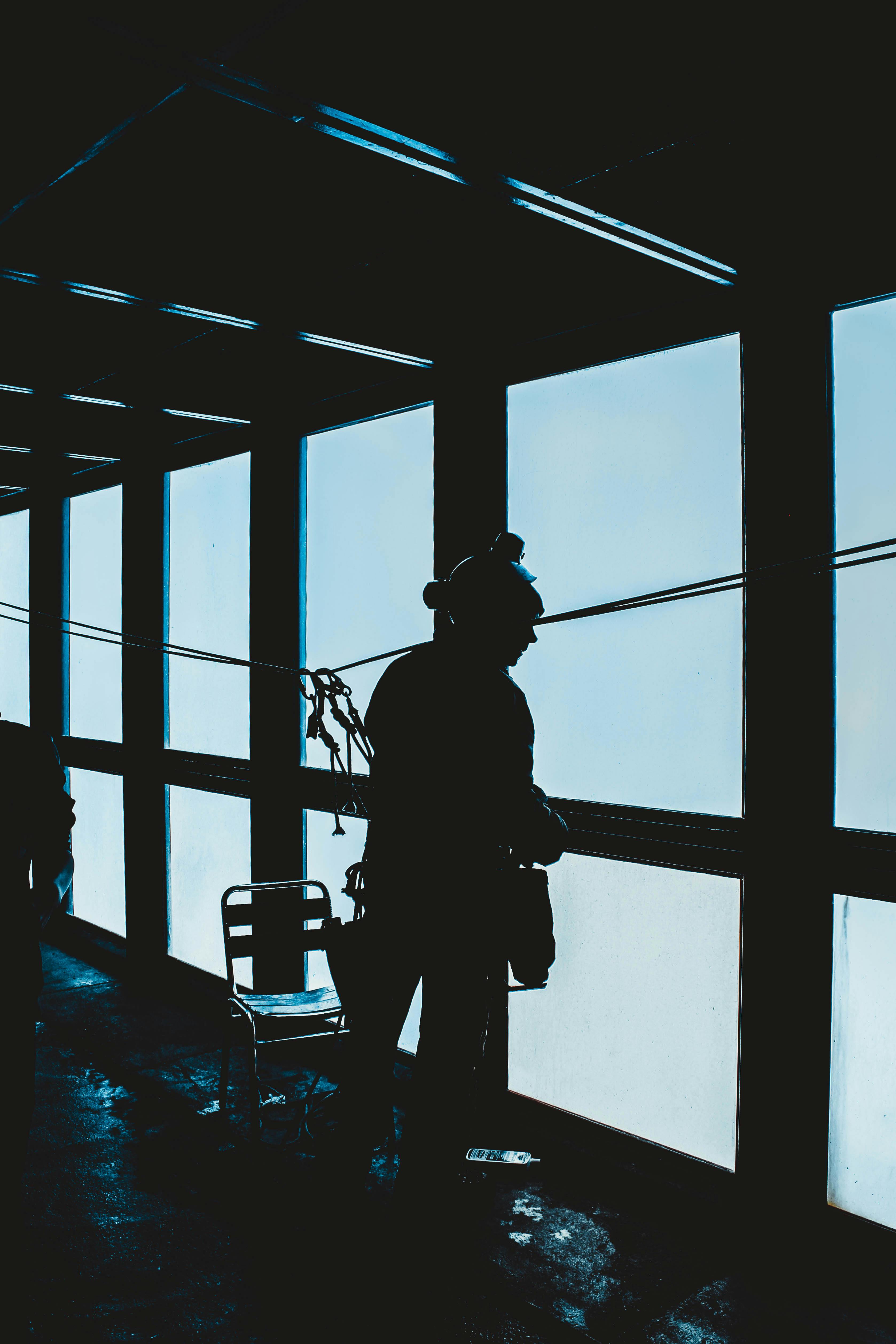 Back view silhouette of expedition member wearing alpinist outerwear standing near big window in observation room and looking out