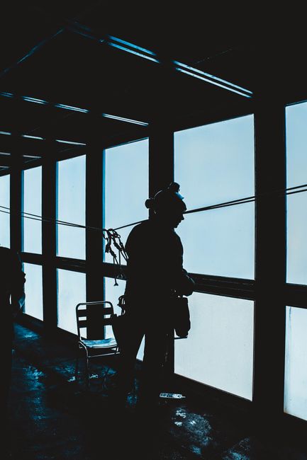 Back view silhouette of expedition member wearing alpinist outerwear standing near big window in observation room and looking out