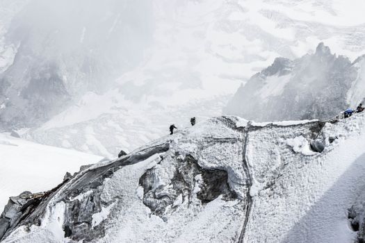 Scenery of distant alpinists climbing on rough massive mountain slope covered with snow against spacious frozen mountainous valley on cold winter day
