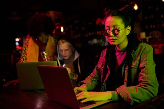 Three diverse adults collaborating on laptops in a dimly lit bar setting.