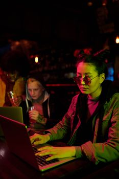 Group of diverse young adults using laptops in a dimly lit bar with vibrant lighting.