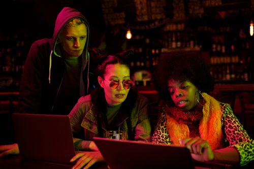 Free Three friends collaborating over laptops in a dimly lit pub setting, showcasing diversity and teamwork. Stock Photo