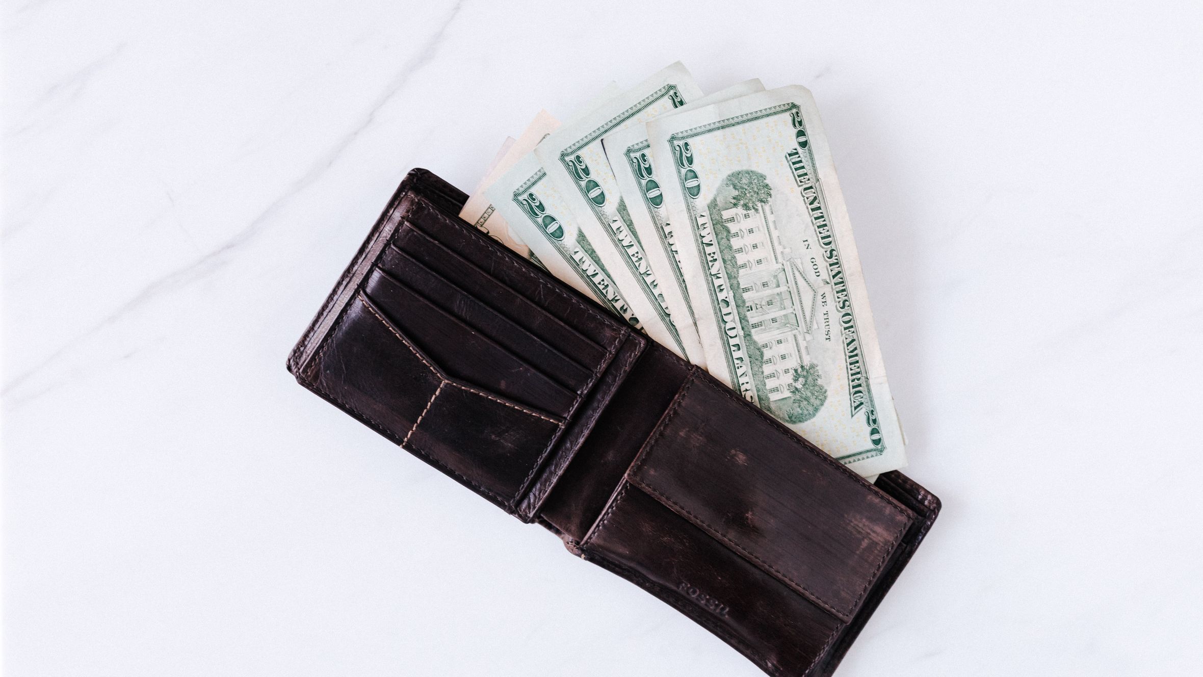From above of black leather wallet with pockets filled with heap of dollar banknotes on white table