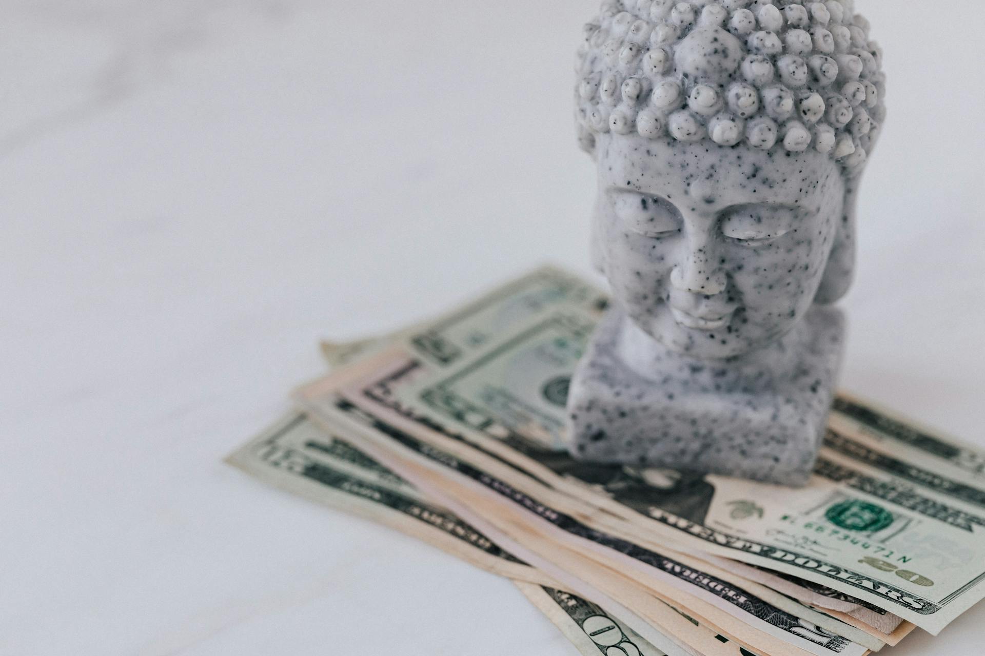 Marble decorative bust of Buddha composed on heap of American dollars on white surface