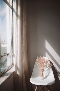A serene minimalist interior with sunlight casting shadows on a chair and dried flowers in a vase.