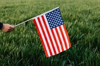 Crop faceless person holding spiky stick with American flag representing stripes and stars of bright colors on field with growing grass in daylight