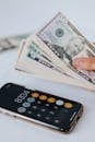 From above of crop anonymous person holding heap of American dollar near cellphone with calculator application representing numbers