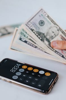 From above of crop anonymous person holding heap of American dollar near cellphone with calculator application representing numbers