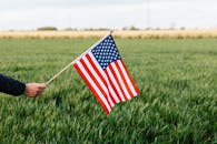 Crop unrecognizable person holding colorful flag of America with stars and stripes on lush green lawn under cloudy sky in daylight