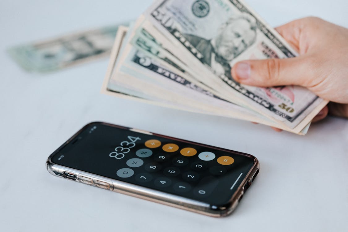 Free Calculator application on mobile phone screen on white surface near crop anonymous person holding pile of dollars Stock Photo