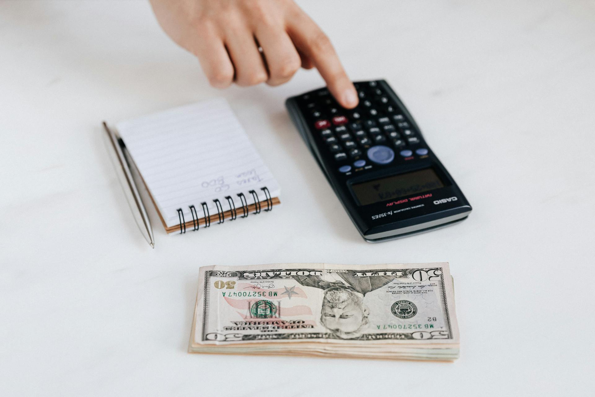 Top view of crop faceless economist touching calculator button while counting income near heap of paper bills and small notebook with pen