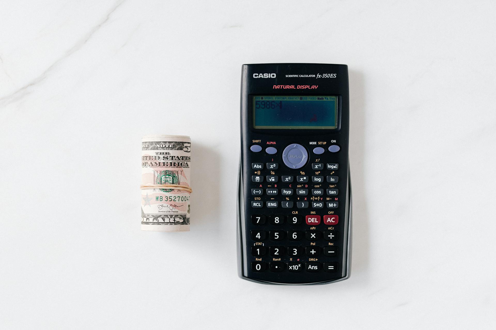 Top view of calculator with plastic multicolored buttons and display near roll of dollar banknotes tied with rubber band on white surface