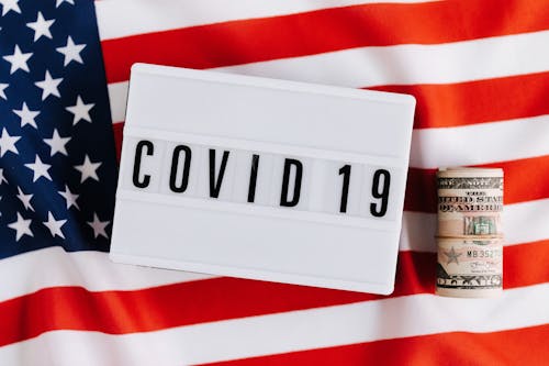 From above of plastic signboard with COVID 19 inscription on flag of USA and roll of paper money during financial crisis