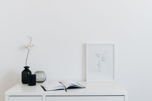 Free A minimalist setup featuring a desk with black vase, open book, and line art frame against a white background. Stock Photo