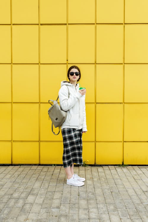 Full body side view of woman in sunglasses and warm wear holding hot drink while standing against yellow wall