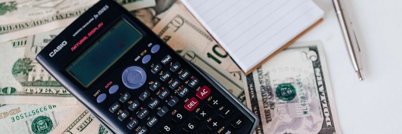 From above electronic calculator and notepad placed over United States dollar bills together with metallic pen for budget planning and calculation