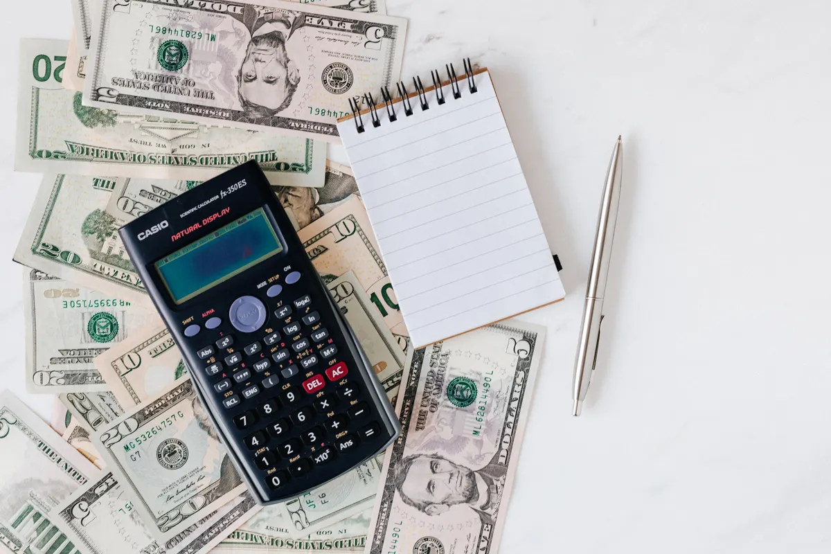A calculator and notepad resting on a stack of US dollar bills, representing an itemized monthly bill