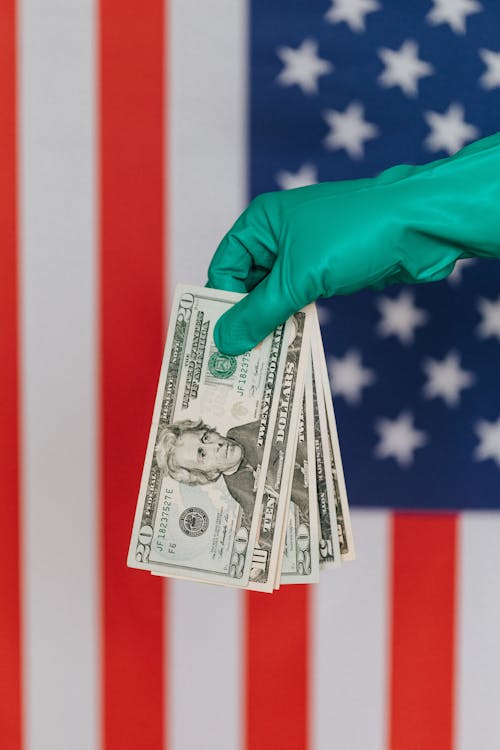 Unrecognizable person wearing green latex glove showing heap of USA banknotes on blurred background of national flag of United States