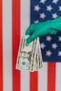 Unrecognizable person wearing green latex glove showing heap of USA banknotes on blurred background of national flag of United States