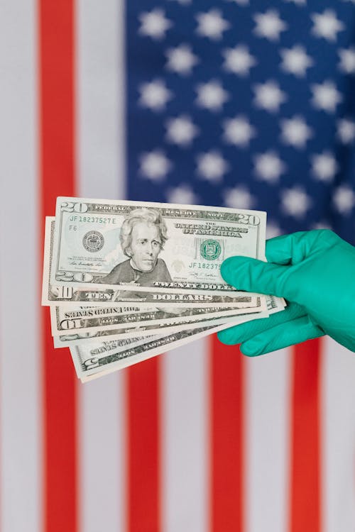 Free Unrecognizable person in medical disposable gloves demonstrating paper banknotes against multicolored American flag on background Stock Photo