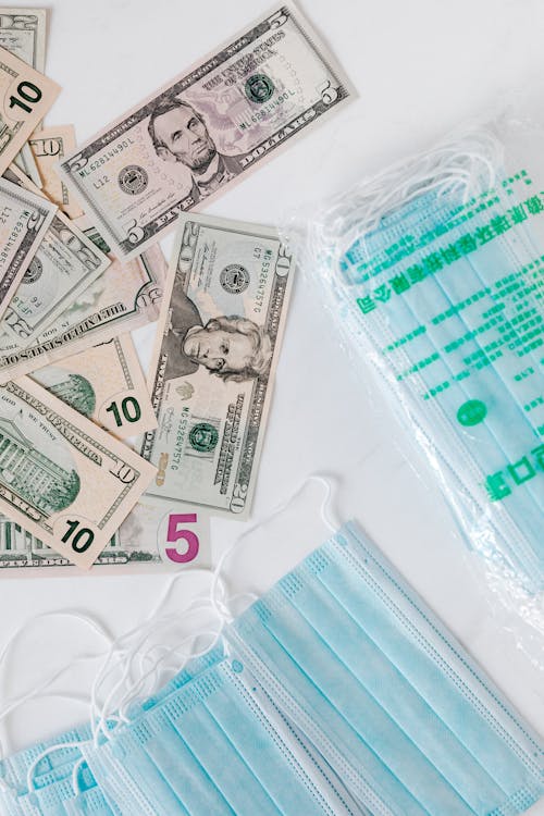 Top view composition of disposable medical masks placed on white table near scattered randomly various paper banknotes of American dollars