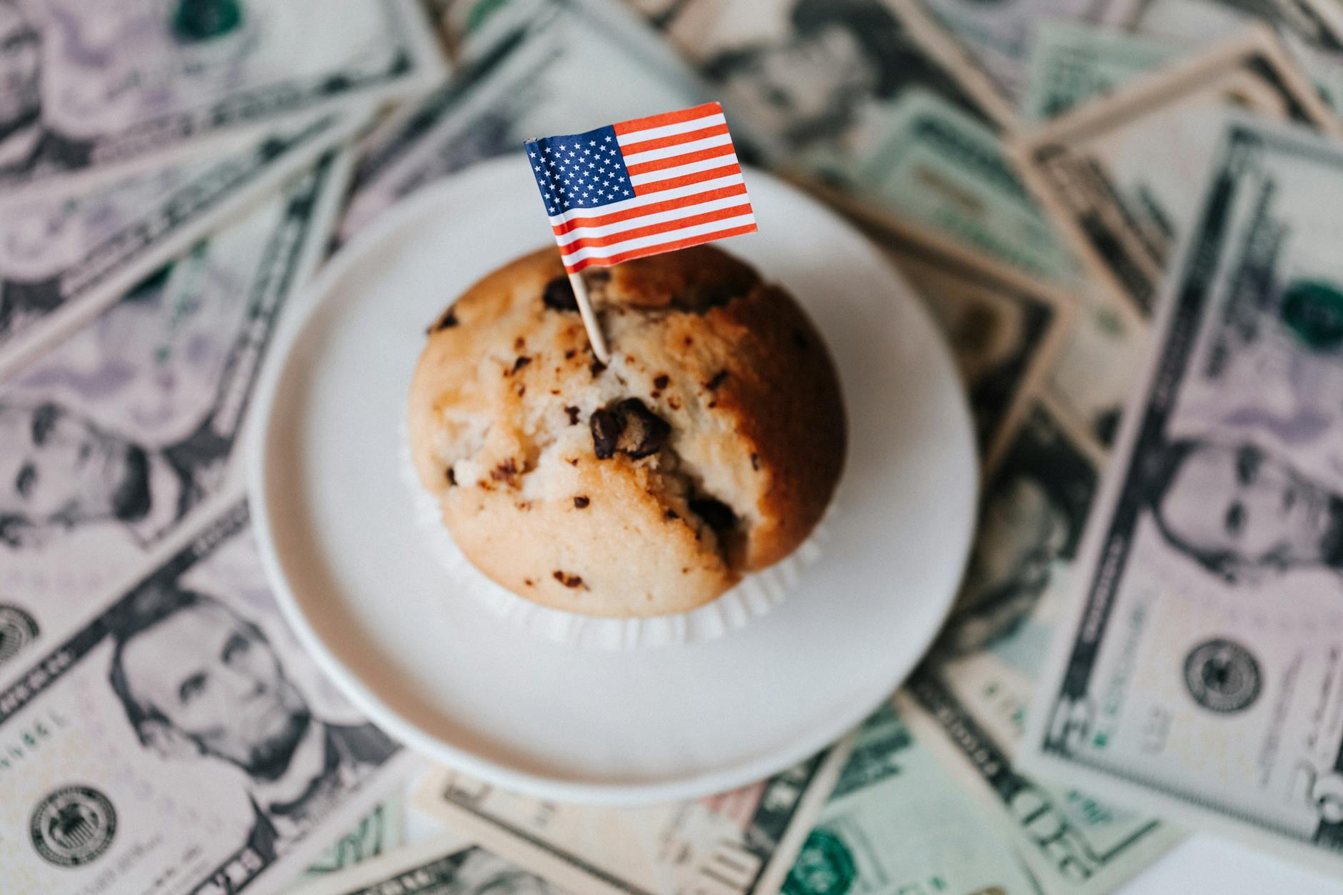 Muffin topped with American flag surrounded by dollar bills, symbolizing wealth and patriotism.