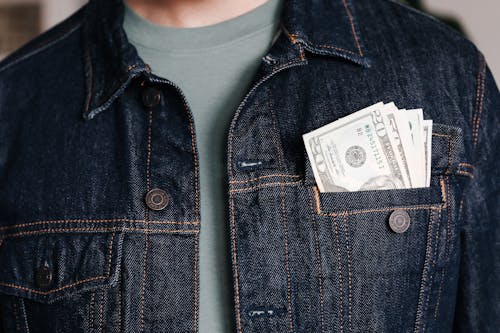 Crop unrecognizable male in casual outfit standing with different nominal pars of dollar banknotes in pocket of jeans jacket