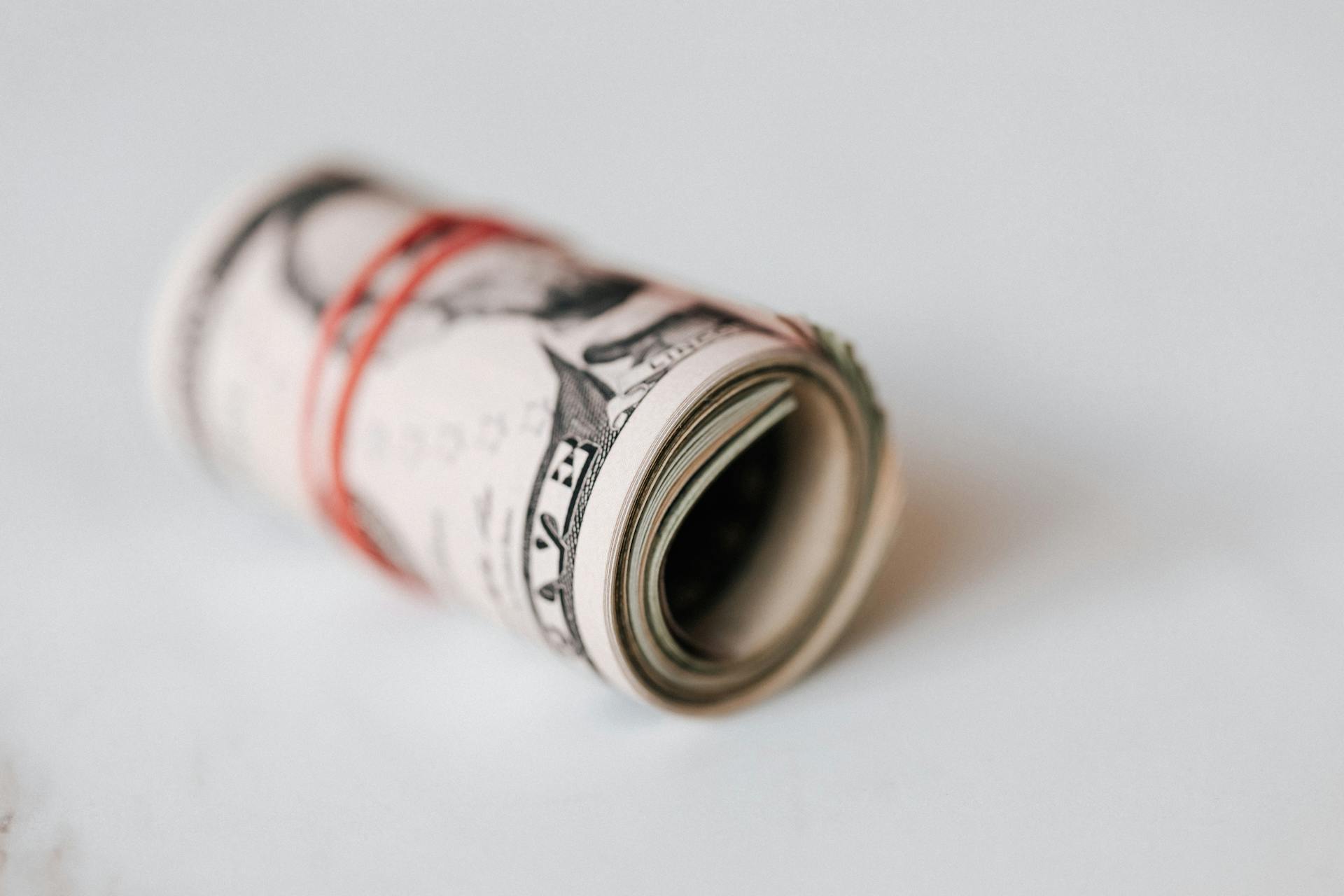 High angle roll of five dollar bills on white table tightened with red rubber band